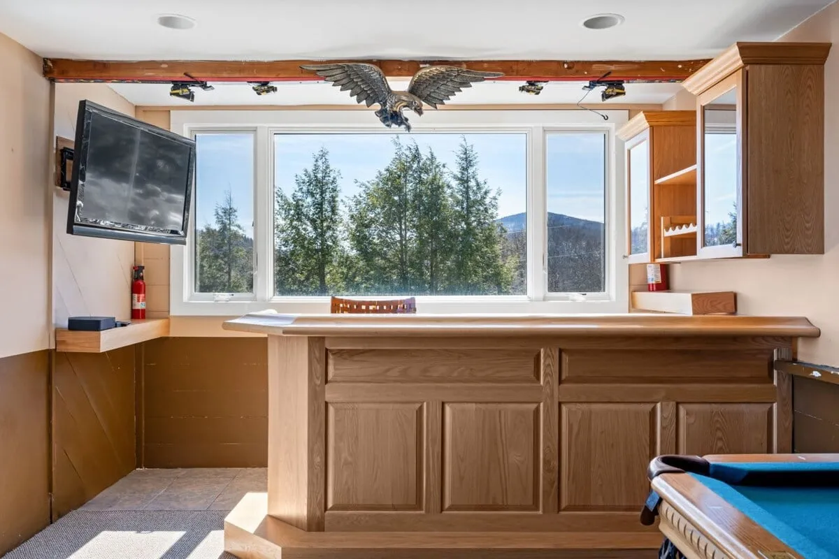 A stylish entertainment suite featuring a wooden bar counter, large window with a scenic mountain view, eagle decor, and a pool table. The image captures a moment of socializing, with guests enjoying drinks at the bar, playing pool, and taking in the view.