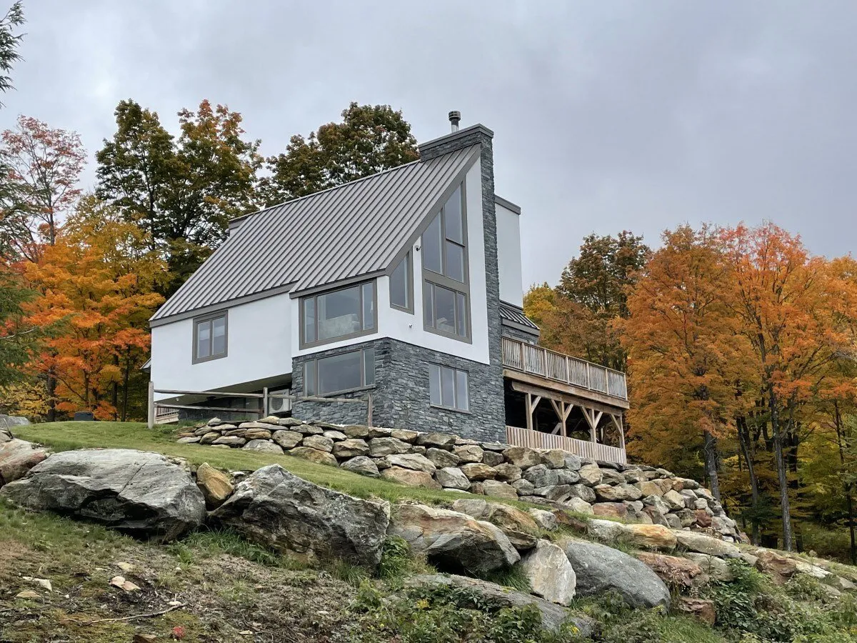 A wide exterior view of The Matterhorn House, showcasing its modern architecture, natural stone facade, and spacious deck area surrounded by vibrant autumn foliage. The deck is visible as an inviting space for outdoor gatherings, set against a beautiful natural backdrop.
