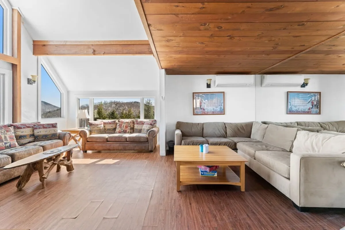 This image features a warm, inviting living room with large windows framing stunning mountain views. Comfortable seating areas, exposed wooden ceiling beams, and a coffee table with books create a cozy space ideal for relaxing or socializing. The arrangement encourages gathering and conversation, and the natural light enhances the atmosphere.