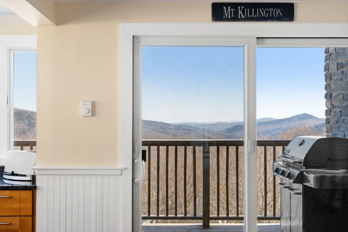 A bright interior shot showing a sliding glass door leading to a deck with a grill and stunning mountain views. This space blends indoor comfort with outdoor access, suggesting opportunities for barbecues and leisurely enjoyment of the landscape.