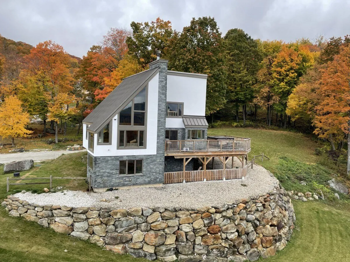 An inviting exterior shot featuring the Matterhorn House with its modern architectural design, surrounded by vibrant autumn foliage. The stone retaining wall and the spacious deck area suggest seamless transitions between the home and nature. The trees display a spectrum of seasonal colors, creating a quintessential Vermont landscape that beckons outdoor adventure.