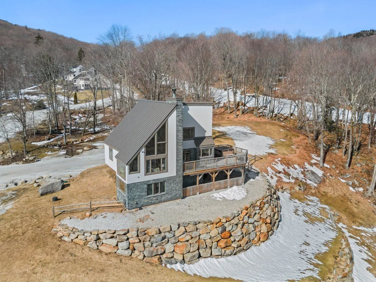 A wide view of the modern house exterior highlights the deck area and stone wall, set against a backdrop of lush trees. The focus is on the outdoor living spaces, suggesting opportunities for gatherings or quiet moments after mountain adventures.