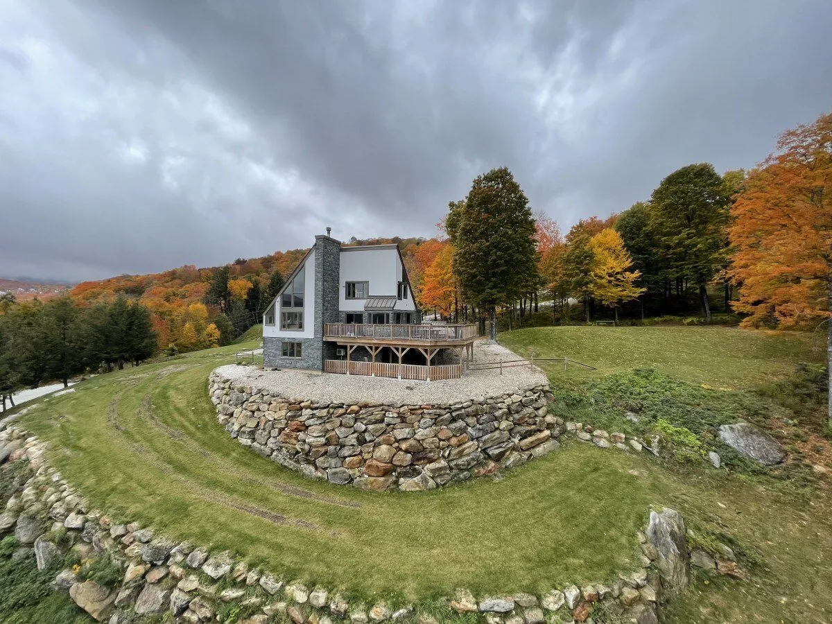 This exterior shot displays the house nestled among mature trees, with a welcoming stone wall and a spacious porch perfect for outdoor relaxation. The tranquil natural surroundings are visible, emphasizing connection to the outdoors and peaceful ambiance.