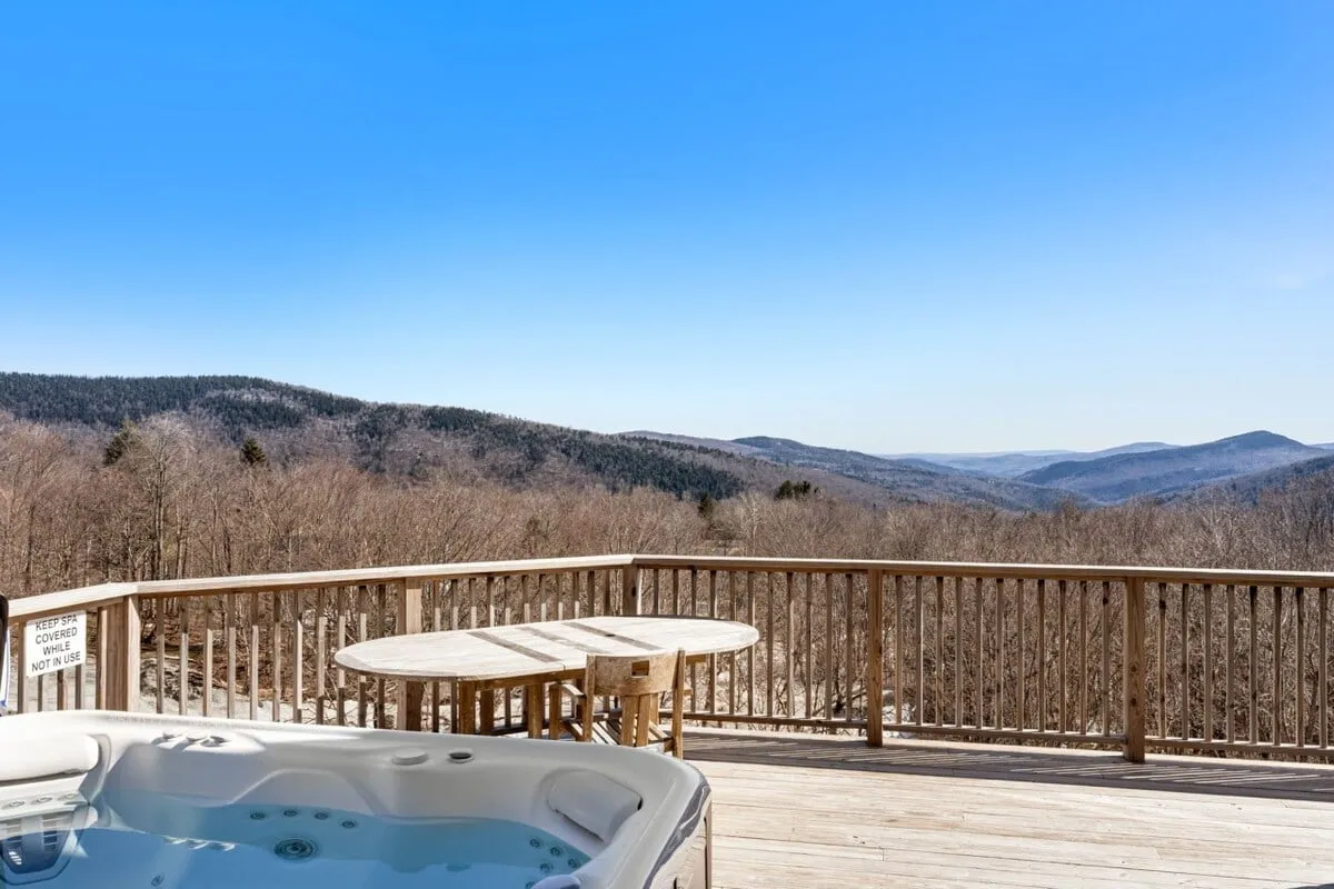 A serene exterior image showing a bubbling hot tub and a wooden table on a deck, overlooking mountain views. The scene blends relaxation with the natural setting, suggesting the perfect post-adventure wind-down while enjoying the crisp mountain air or stargazing in the evening.