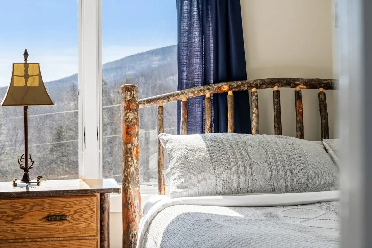 A warmly decorated bedroom with a rustic wooden bed frame, dressed in plush, soft-textured bedding. A unique table lamp sits atop a nightstand, and a large window frames a tranquil mountain view, inviting natural light into the serene space. The overall ambiance is calm and restful, ideal for unwinding after adventurous days.