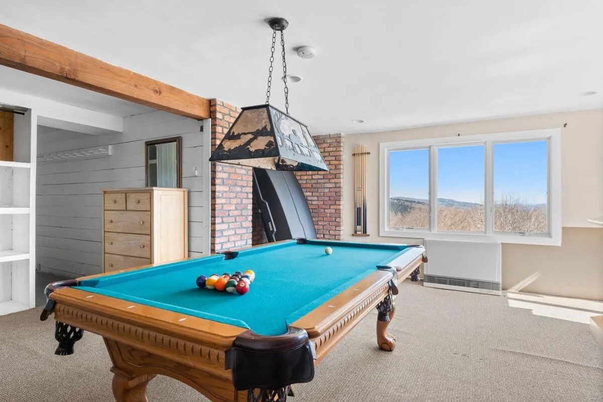 A bright, recreational interior with a pool table set against a brick wall and large windows that flood the room with natural light. The space feels relaxed and social, with areas for playing billiards and lounging with friends.