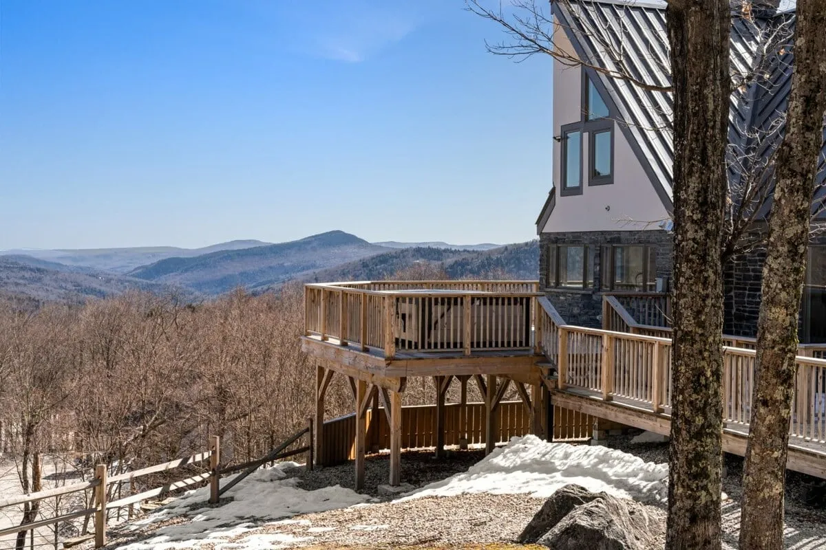 A wide deck extends from the modern house, framed by lush trees and a breathtaking mountain vista. Outdoor seating is visible, offering a peaceful setting for gatherings, stargazing, or simply enjoying the fresh mountain air.