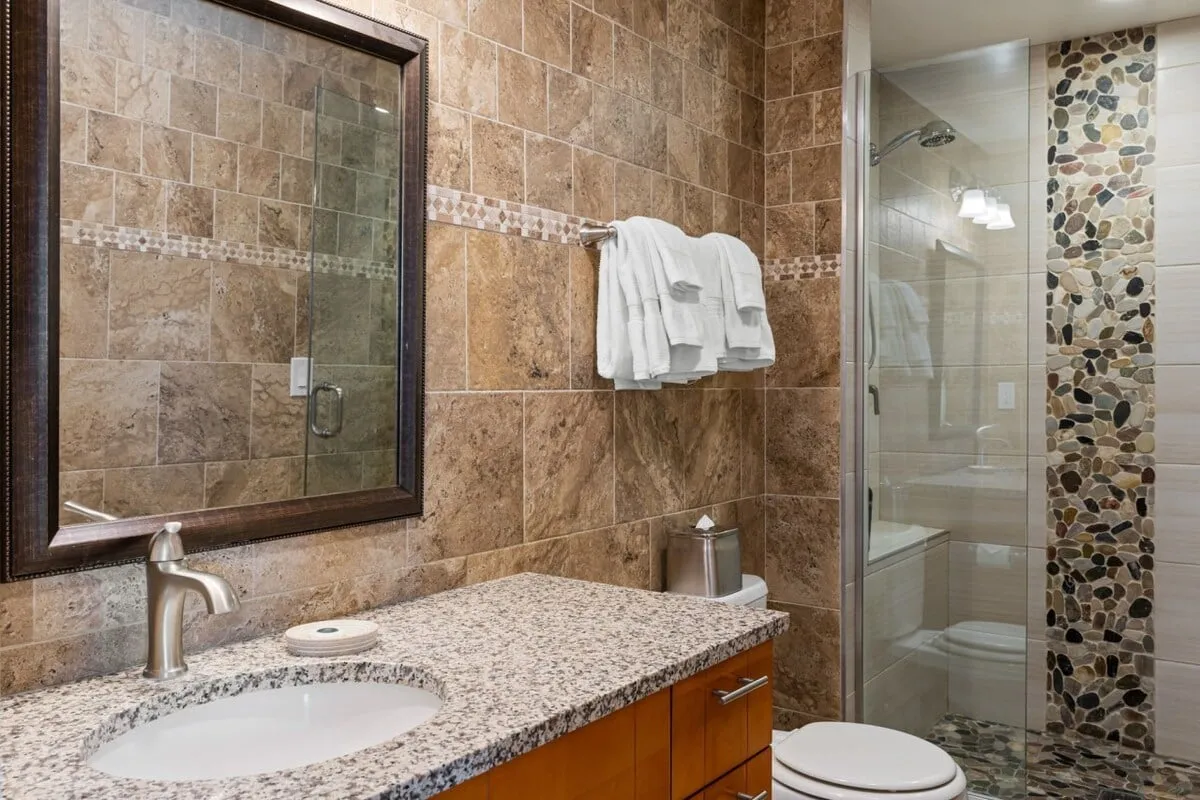 A luxurious, modern bathroom featuring a granite countertop, large mirror, tiled shower stall, and fluffy white towels. The design is upscale and serene, inviting relaxation and making self-care routines a pleasure after a day of adventure.