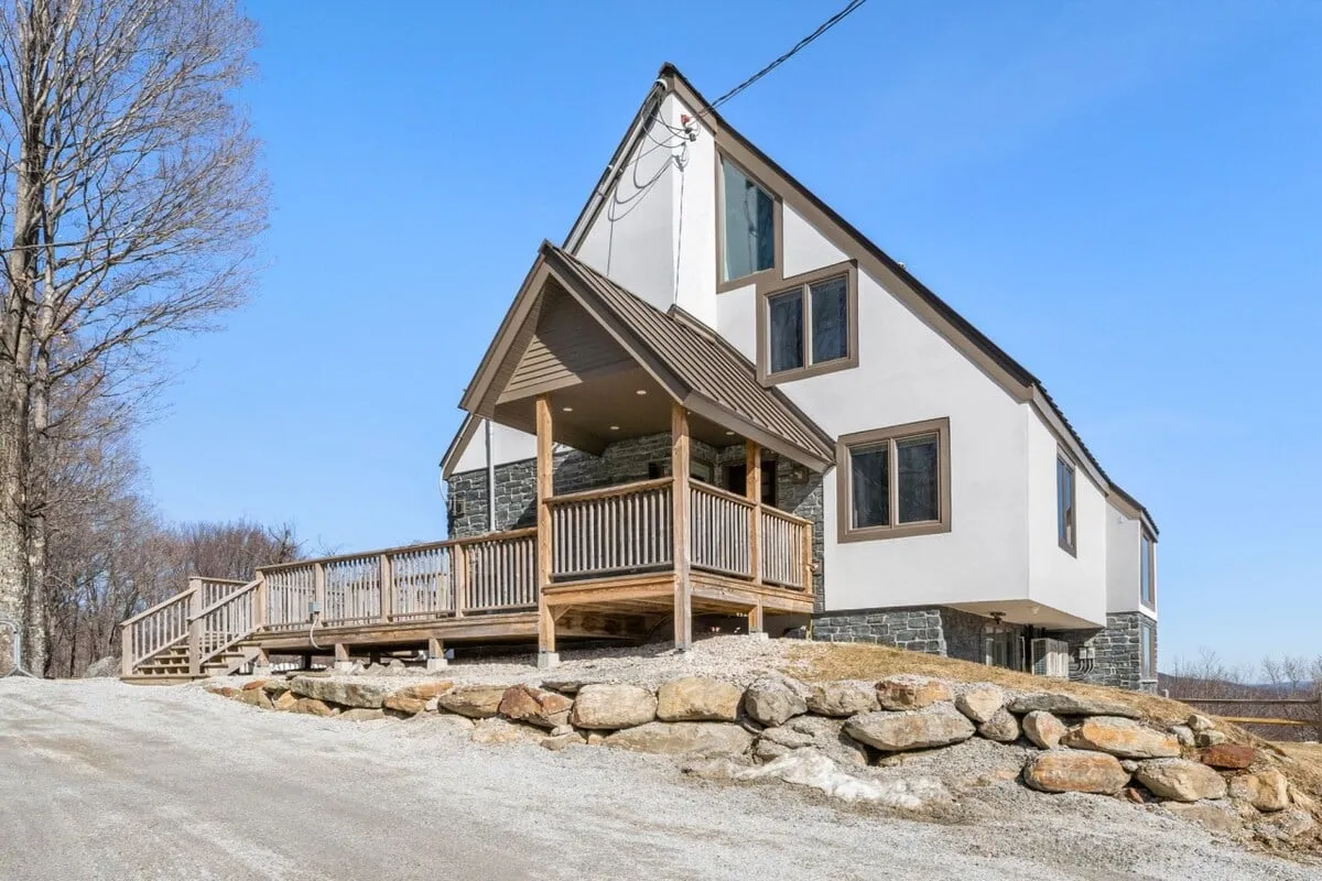 An inviting exterior shot featuring the house's front porch with a stone facade, large windows, and cozy seating area. The porch looks ready for relaxation and group conversations, framed by natural surroundings.