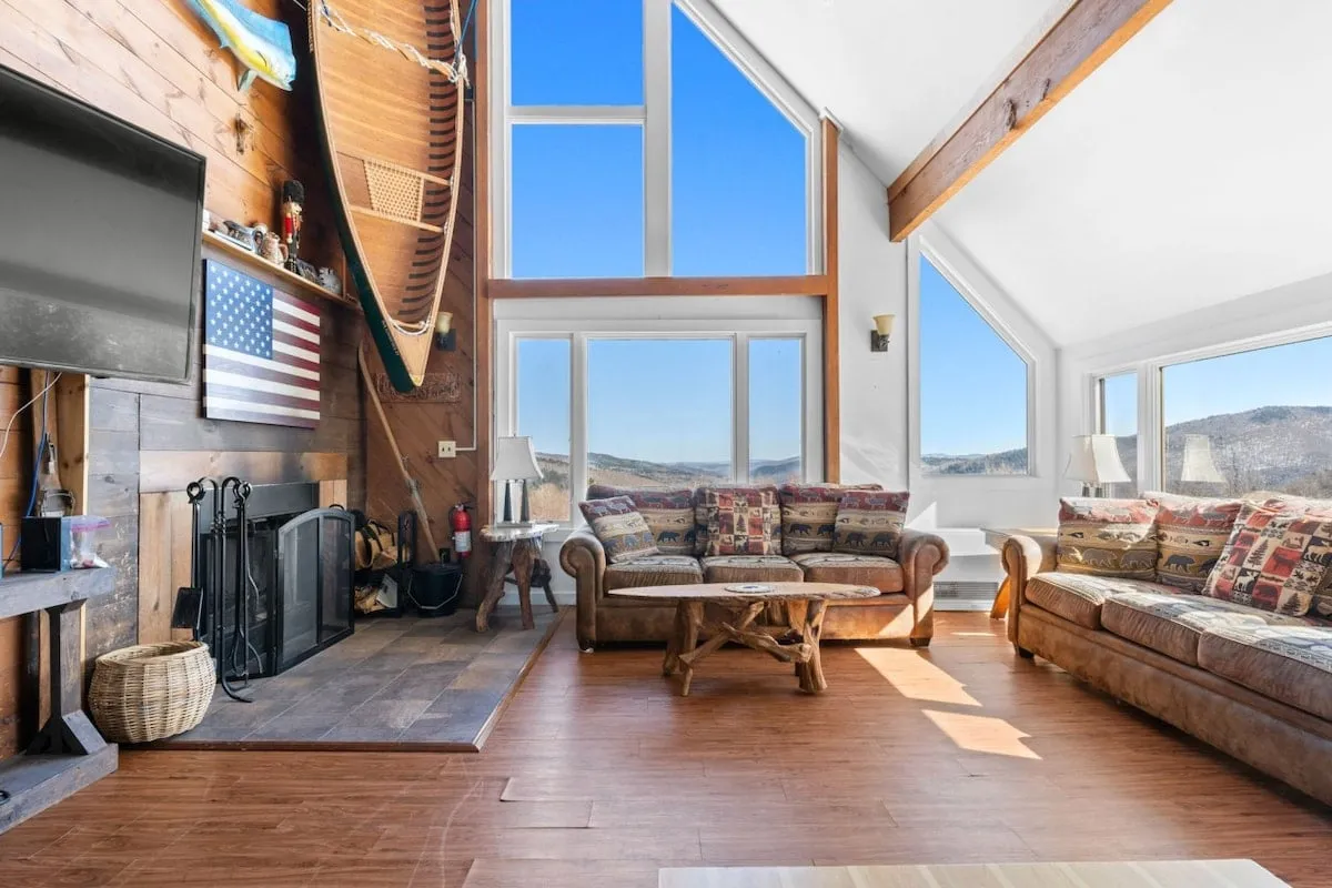 The hero image showcases a spacious, inviting living room with expansive windows framing breathtaking mountain views. The interior features warm, rustic wooden furniture, a decorative canoe mounted on the wall, and a stone fireplace beneath a mounted television, creating a cozy, nostalgic ambiance perfect for gathering with family and friends.