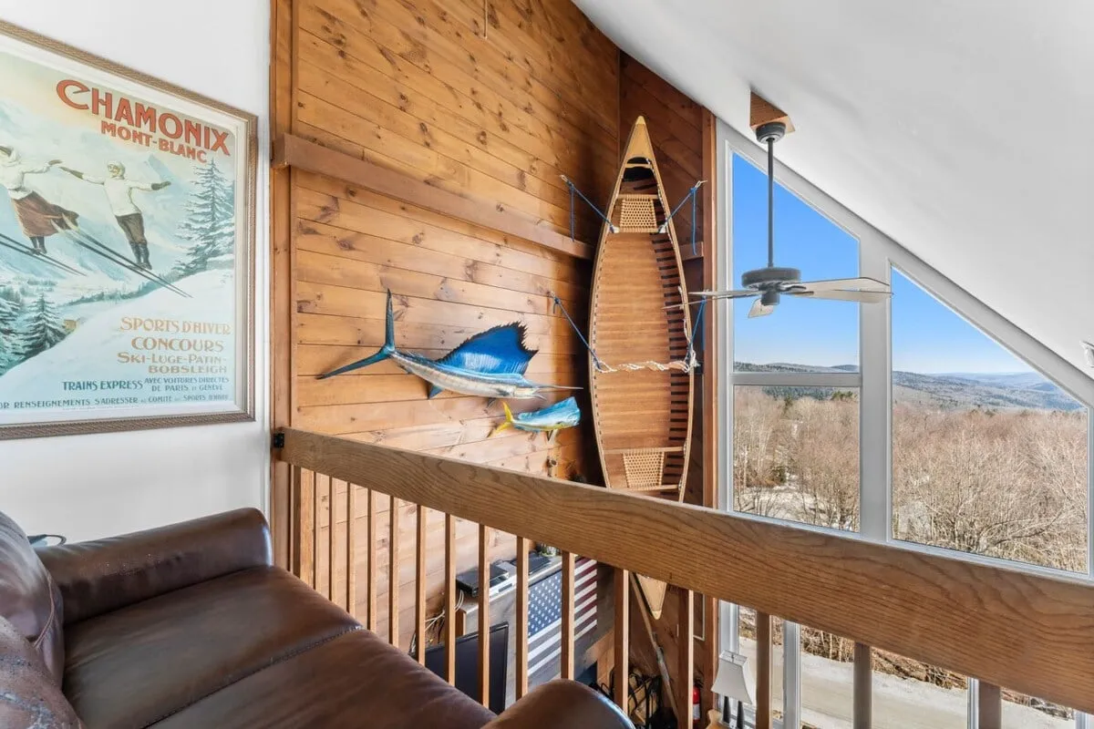 A cozy living space featuring a prominent kayak, fish decorations, a wooden wall, vintage ski poster, and large windows looking out over the mountains. The presence of the kayak and decor nods to outdoor water activities, while the windows visually connect the interior with the surrounding nature.