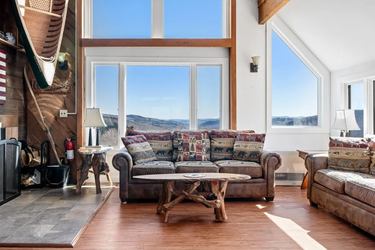 A spacious, open-plan living area filled with rustic wooden furniture, colorful cushions, a canoe wall decoration, and a prominent fireplace. Large windows offer breathtaking mountain vistas, making the space feel connected to nature while fostering warmth and togetherness.
