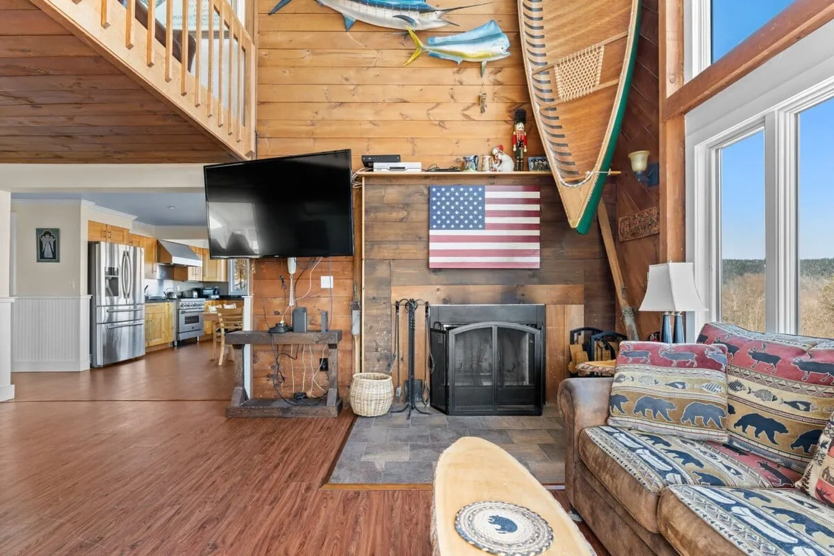 A warm living room with a large American flag above a stone fireplace, plush sofa with wildlife print, wooden walls, and expansive windows letting in natural light. The setting invites relaxation and social gatherings, hinting at the après-ski comfort the home offers after a night on the town.