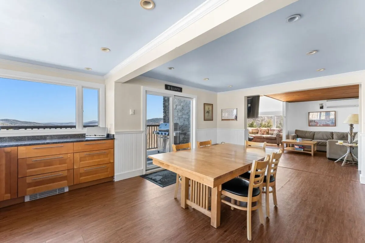 This image highlights the dining area, centered around a large dining table with seating for several people. The space is brightened by large windows that offer scenic views, and the adjacent kitchen cabinetry suggests seamless flow between cooking and dining.